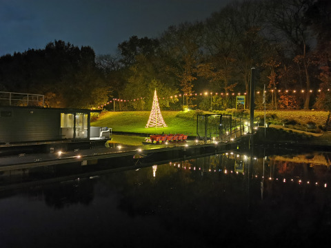 Árbol de Navidad iluminado y luces en una colina, visto desde Houseboat Marina Mookerplas por la noche, Países Bajos.