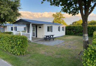 Mobilhome met blauwe dak en veranda, picknicktafel op terras, groene haag op vakantiepark Duinhoeve.