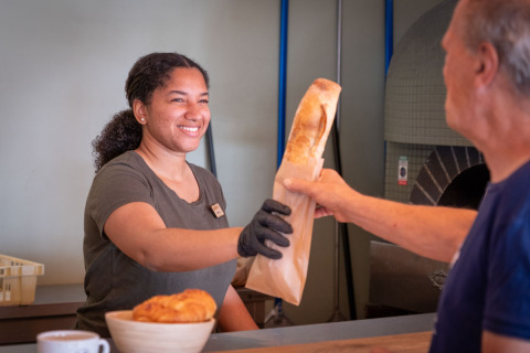 Eine Frau hinter der Theke lächelt und reicht einem Kunden ein Baguette im Huttopia Caminos de Galicia.