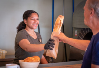 Una mujer sonríe mientras entrega una baguette a un cliente en Huttopia Caminos de Galicia, Galicia, España.