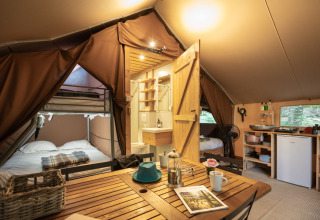 Interior of a cozy Trappeur safari tent featuring beds, a small kitchen area, and a wooden dining table.