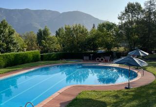 Piscina all'aperto con bordo in legno e ombrelloni, circondata da alberi e montagne sullo sfondo.