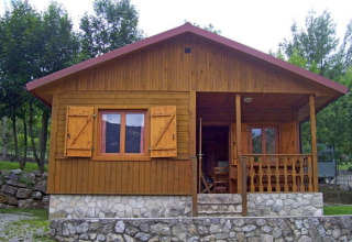Wooden chalet with stone base and small front porch at Huttopia Barcelona Pirineos in Spain, surrounded by trees.