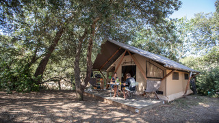 Dos personas se sientan frente a una tienda safari Trappeur en el bosque, rodeadas de árboles altos.