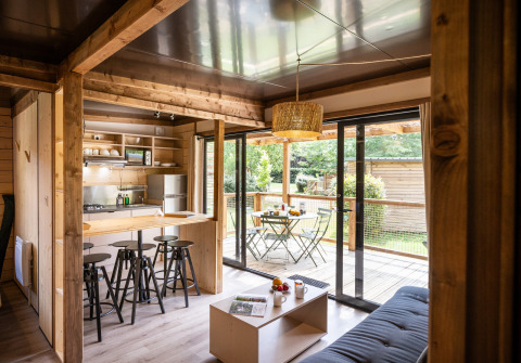 Modern lodge-interieur met keuken, barkrukken, zithoek en zicht op terras en tuin via glazen schuifdeuren.