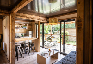 Modern lodge-interieur met keuken, barkrukken, zithoek en zicht op terras en tuin via glazen schuifdeuren.