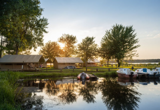 Puesta de sol en la orilla del lago de Huttopia Lac d'Aureilhan - Mimizan, con tiendas, árboles y botes.