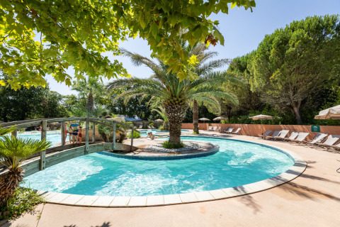 Pool mit Palmeninsel und Brücke im Huttopia Lac d'Aureilhan - Mimizan Feriendorf in Frankreich, umgeben von Liegen.