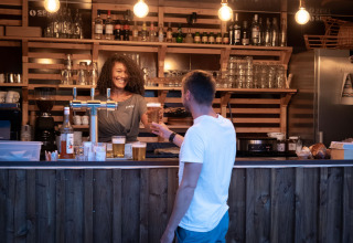 Man in white shirt receiving draft beer from smiling bartender at the wooden bar in Huttopia Lac d'Aureilhan, France.