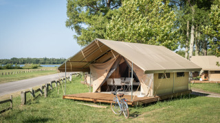 Tienda safari llamada Classic Tent con terraza de madera y bicicleta, ubicada en un entorno natural verde.