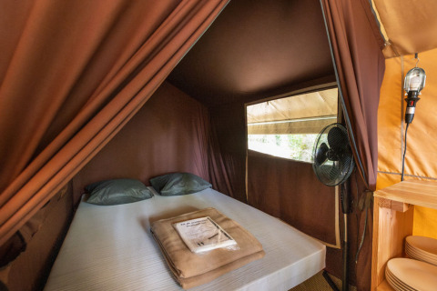 Zona notte in tenda safari classica con letto matrimoniale, ventilatore e finestra a Huttopia Lac d'Aureilhan.