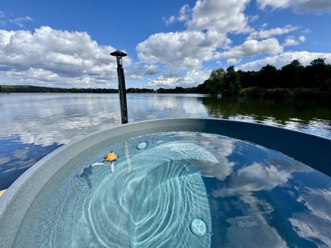 Vista de un lago tranquilo desde un alojamiento de glamping de lujo con jacuzzi redondo al aire libre y cielo azul.