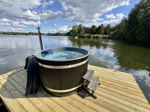 Holzbefeuerte Badetonne auf einer Holzterrasse am See, umgeben von Natur und Bäumen.