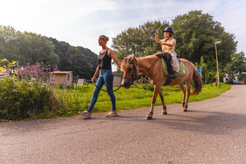 Woman leads a pony with a child riding through Vakantiepark IJsselhof, a holiday park in the Netherlands.