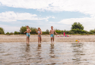Three children play by the water's edge on a sandy beach near Andijk, Netherlands, on a sunny day.