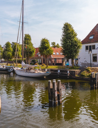 Zeilboten aangemeerd langs een kanaal nabij Andijk in Noord-Holland, met bomen en gezellige gebouwen.