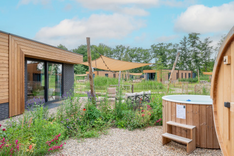 Moderne lodge med træbeklædning, udendørs spabad, blomsterhave og solsejl under en blå himmel.