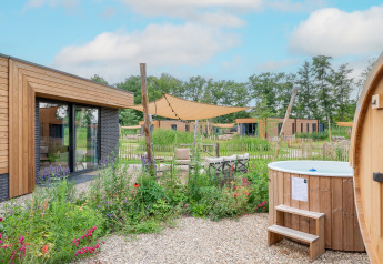 Lodge moderno con revestimiento de madera, jacuzzi exterior, jardín de flores y toldos bajo cielo azul.