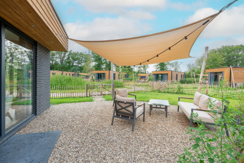Gezellige buitenhoek bij een moderne lodge met zonnedoek, loungezetels en zicht op groene natuur.