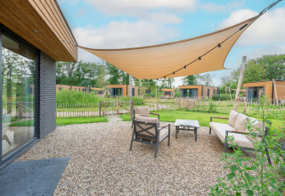 Buiten zithoek bij een moderne lodge met schaduwdoek, comfortabele stoelen en uitzicht op groen.