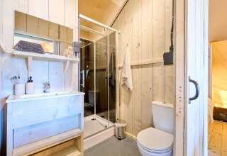 Salle de bain lumineuse dans un hébergement glamping avec murs en bois, douche, lavabo et toilettes.