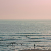 Beach near Aureilhan, Nouvelle-Aquitaine, France with people swimming and surfing under a soft pink sky.