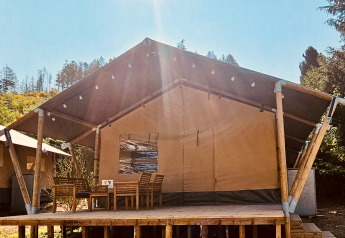 Safaritent met houten veranda, eettafel en stoelen in de zon, omringd door bomen en natuur