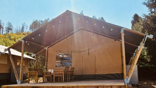Safari-Zelt mit Holzterrasse, Esstisch und Stühlen im Sonnenlicht, umgeben von Bäumen
