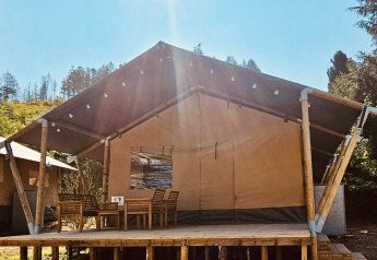 Safari-Zelt mit Holzterrasse, Esstisch und Stühlen im Sonnenlicht, umgeben von Bäumen