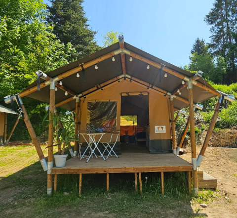 Safari tent with a wooden deck, string lights, and outdoor furniture, surrounded by greenery on a sunny day.