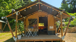 Safari tent with a wooden deck, string lights, and outdoor furniture, surrounded by greenery on a sunny day.