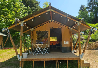 Tenda safari con veranda in legno, luci decorative e arredo da esterno, immersa nel verde in una giornata soleggiata.