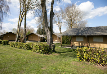 Safaritenten met privé sanitair omringd door hagen, gras en bomen onder een heldere blauwe lucht.
