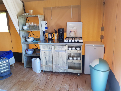 Área de cocina en una tienda safari con estufa de gas, nevera, estantes, utensilios y cubo de basura en suelo de madera.