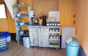 Área de cocina en una tienda safari con estufa de gas, nevera, estantes, utensilios y cubo de basura en suelo de madera.