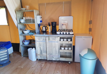 Keukengedeelte in een safaritent met gasvuur, koelkast, rekken, serviesgoed en vuilnisbak op houten vloer.