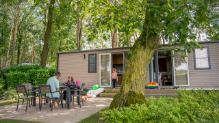 Family relaxing outside at a table near an Extra Luxe Stacaravan mobile home, surrounded by trees.