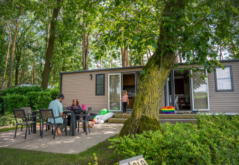 Familie entspannt auf der Terrasse vor einem Extra Luxe Stacaravan Mobilheim zwischen Bäumen.