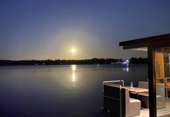 Foto nocturna desde una casa flotante con agua tranquila, luz de luna y comedor exterior junto al lago.