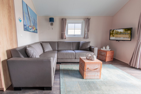 Moderne woonkamer met grijze hoekbank, houten tafeltjes en tv in de Lodge op Vakantiepark Leukermeer, Nederland.