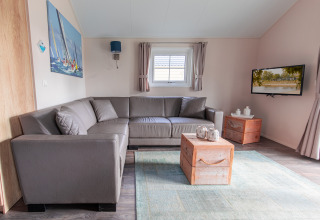 Moderne woonkamer met grijze hoekbank, houten tafeltjes en tv in de Lodge op Vakantiepark Leukermeer, Nederland.