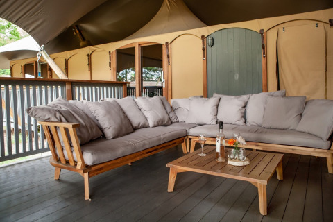 Comfortabele zithoek met houten bank, grijze kussens en lage tafel op het terras bij Sandberghe vakantiepark.