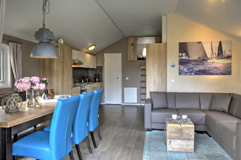 Moderne lodge met blauwe stoelen, grijze hoekbank en zeilfoto in Vakantiepark Leukermeer, Nederland.