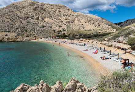 Playa de guijarros con sombrillas y agua cristalina cerca de Krk, Croacia, rodeada de colinas rocosas.