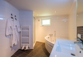 Baño luminoso con bañera de esquina, lavamanos, albornoces y ventana en Wellness Lodge de De Wiltzangh.