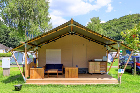 Safari tent without sanitation, furnished with wood on a deck, set on a grassy campsite under blue sky.