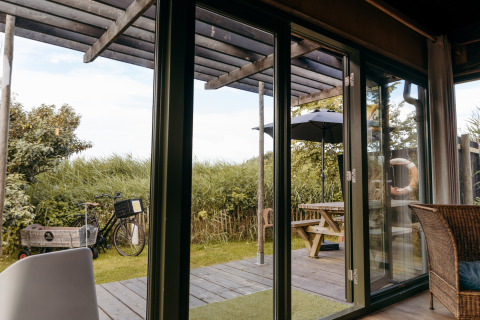 Uitzicht vanuit een lodge met glazen deuren naar terras met picknicktafel, fiets en parasol bij Duynpark Het Zwanenwater.