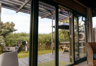 Uitzicht vanuit een lodge met glazen deuren naar terras met picknicktafel, fiets en parasol bij Duynpark Het Zwanenwater.