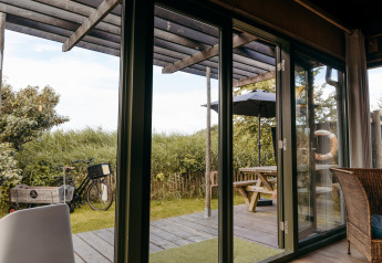Vista desde una cabaña con puertas de cristal hacia una terraza con mesa, sombrilla y bicicleta en Duynpark Het Zwanenwater.
