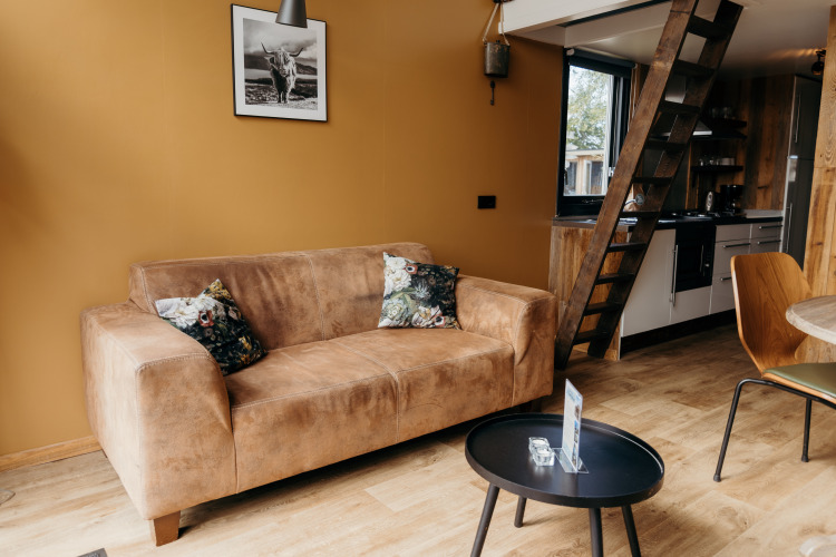Sala de estar en Rescue cottage de Duynpark Het Zwanenwater, con sofá marrón, escalera y cocina.